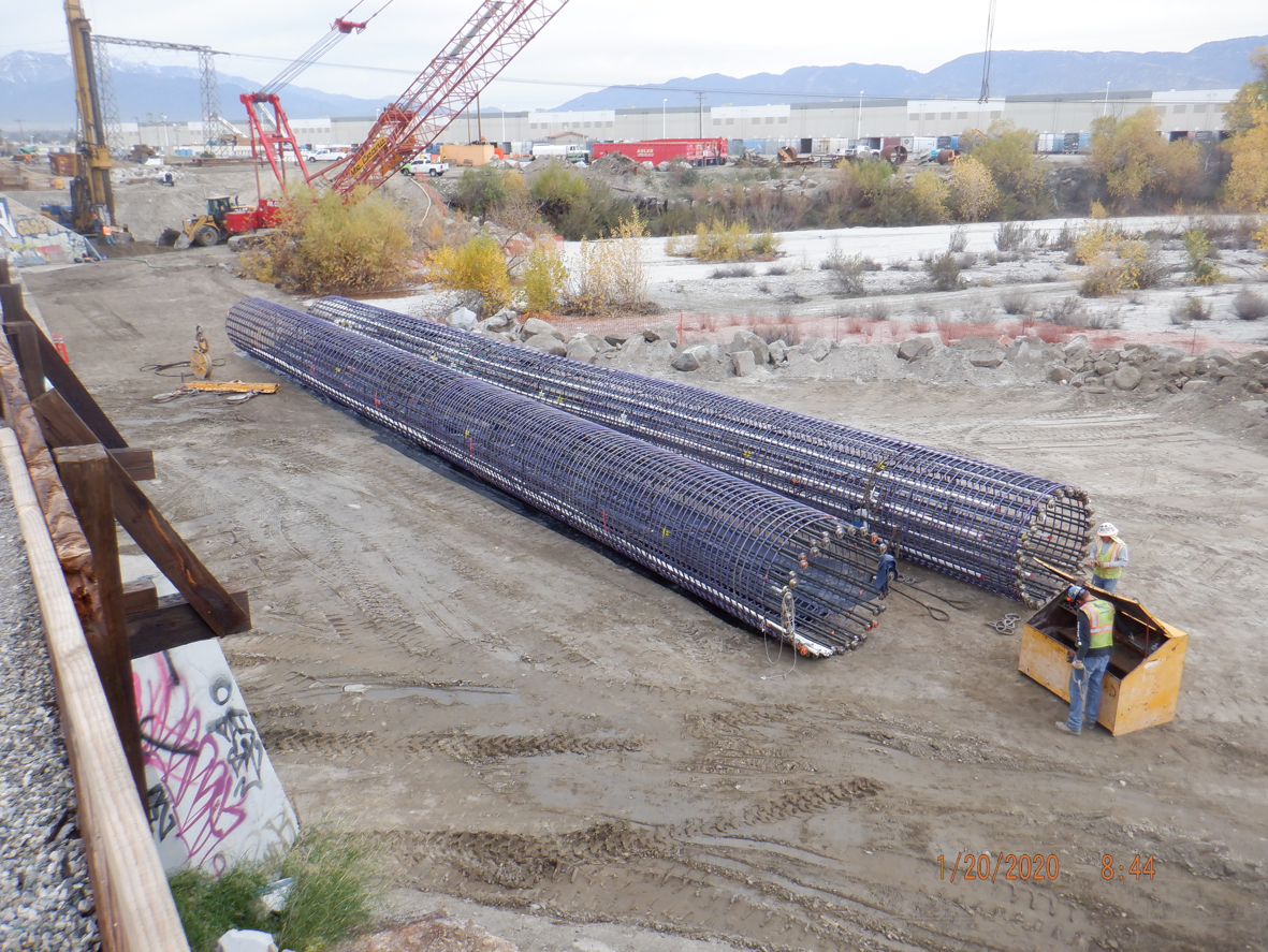 Three large cylindrical steel rebar cages lie on the ground at a construction site, with workers and cranes visible in the background.