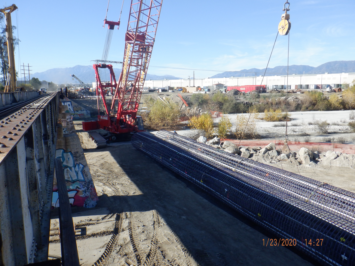 A large red crane and a hook are positioned at a construction site with steel rebar cages, near a railway bridge and industrial buildings, under a clear sky.