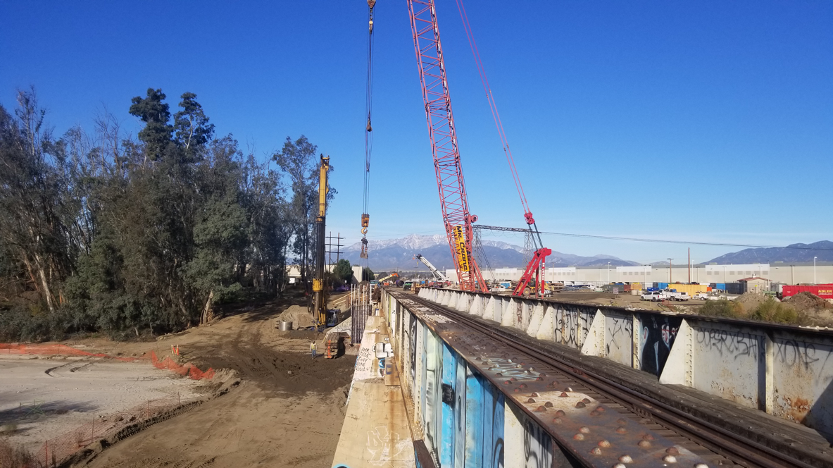 A construction site next to railroad tracks with cranes and machinery in use, trees on the left, industrial buildings on the right, and mountains in the background under a clear blue sky.