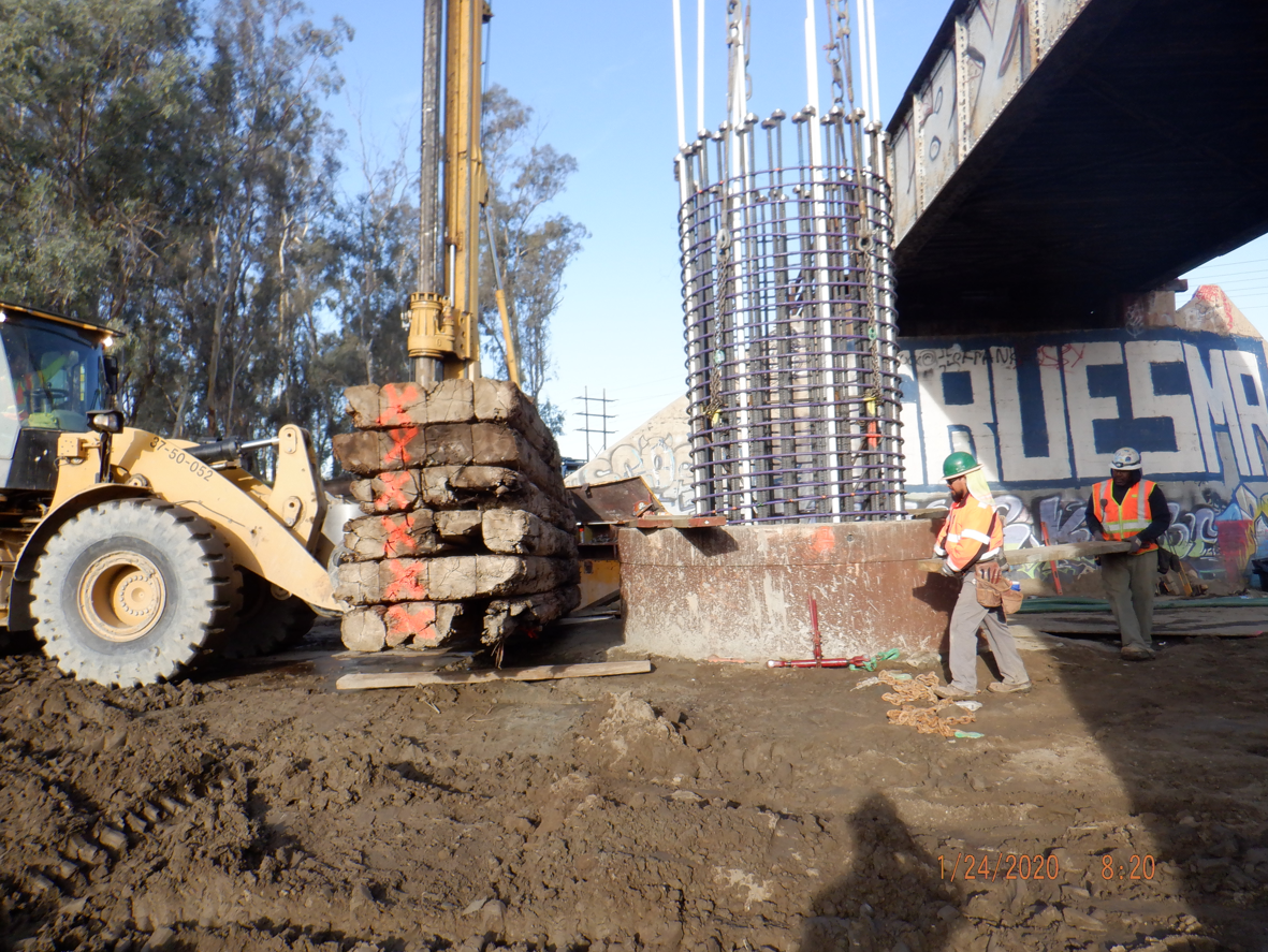 Construction workers operate heavy machinery near a bridge support structure, with rebar columns and graffiti visible, on a muddy worksite.