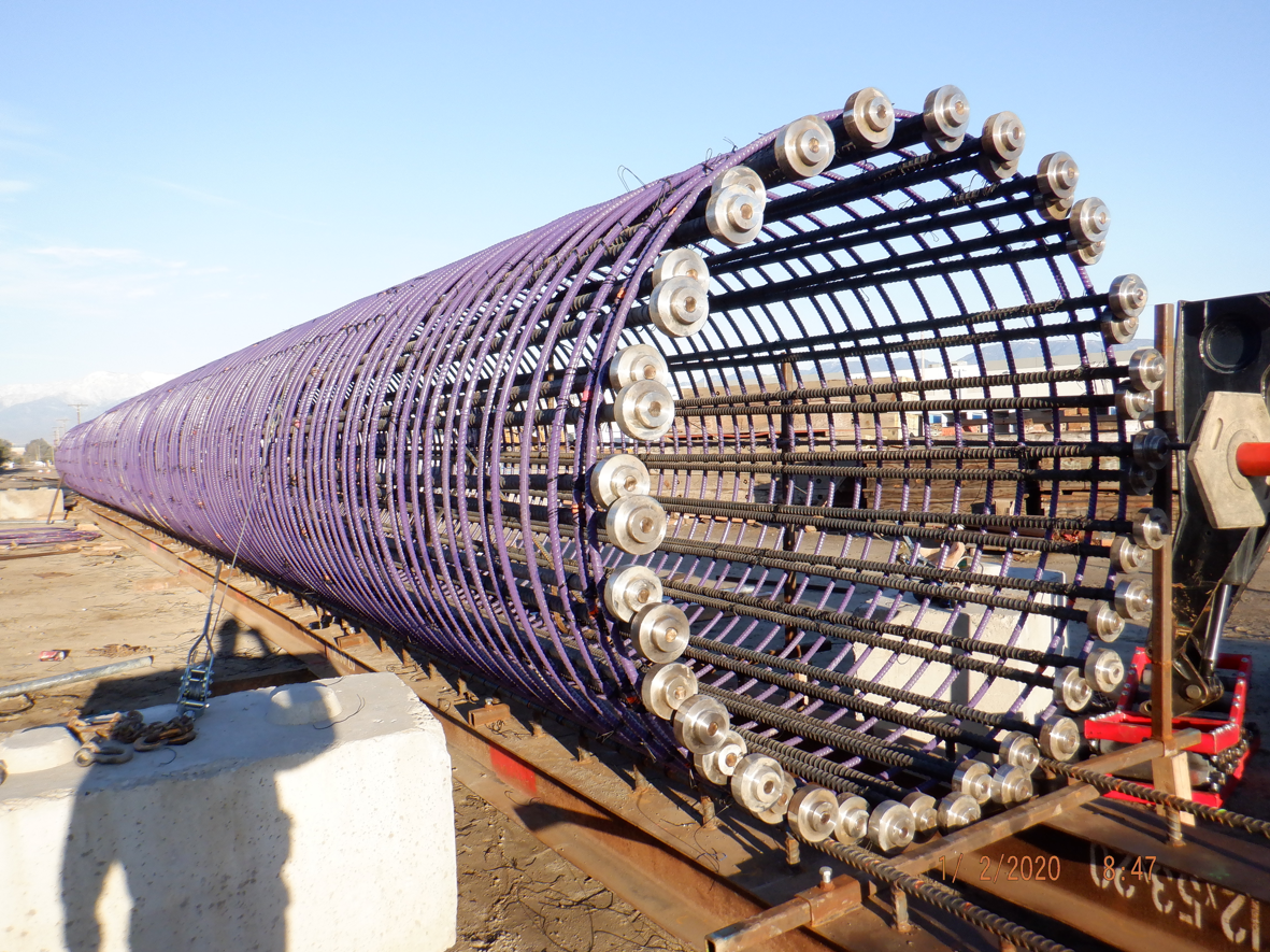 A large cylindrical steel rebar cage with purple-coated rods and metal discs is laid horizontally at an outdoor construction site.