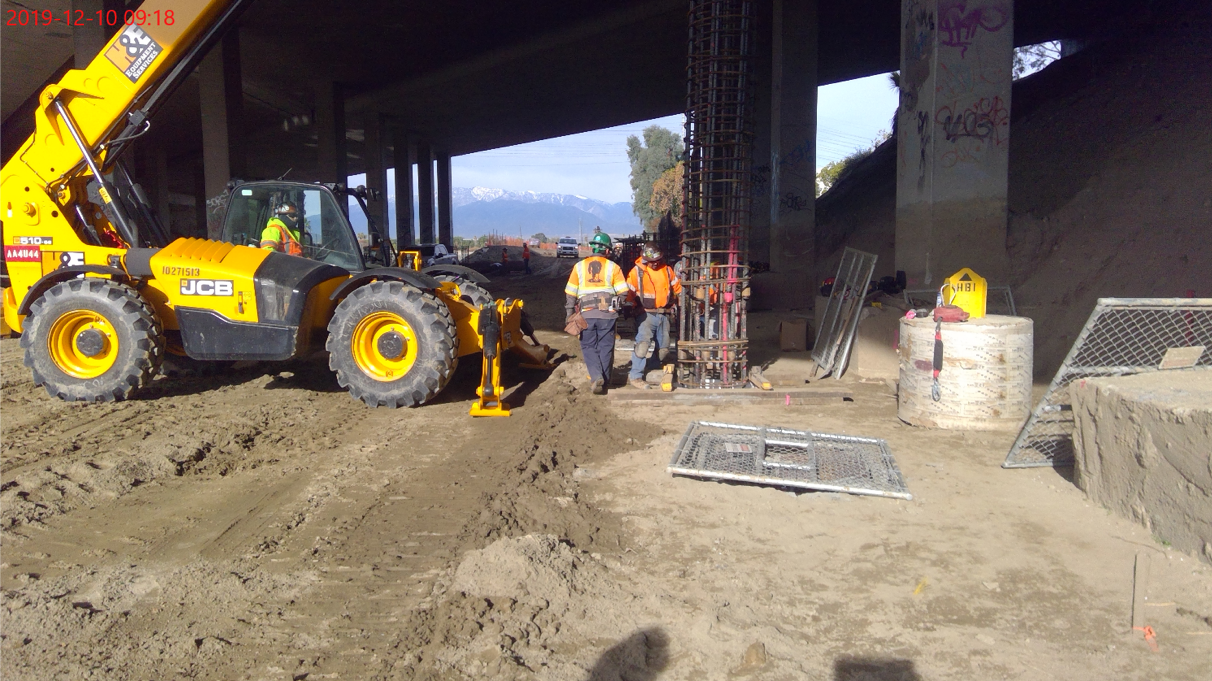 Construction workers operate a yellow JCB telehandler and work near a rebar column under a bridge at a construction site. Dirt and equipment are scattered around.