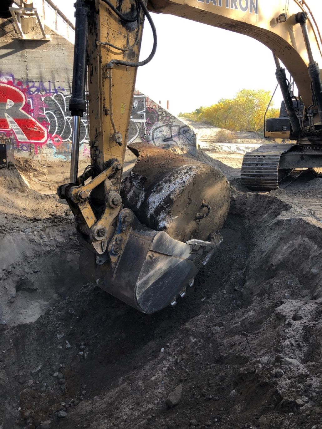 A yellow excavator digging a trench in a dirt area near concrete structures with graffiti.