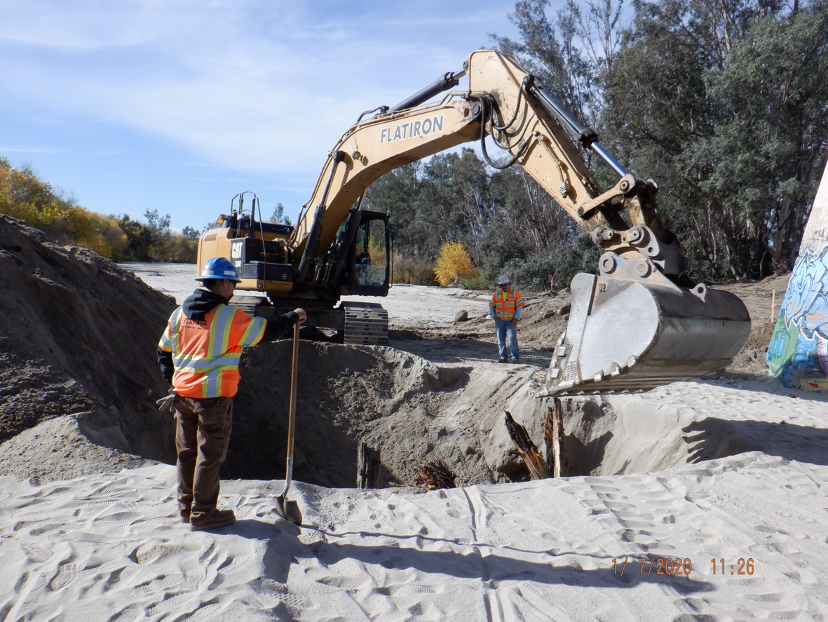 Two construction workers in safety vests and helmets stand near an excavator digging sand at an outdoor site with trees and graffiti in the background.