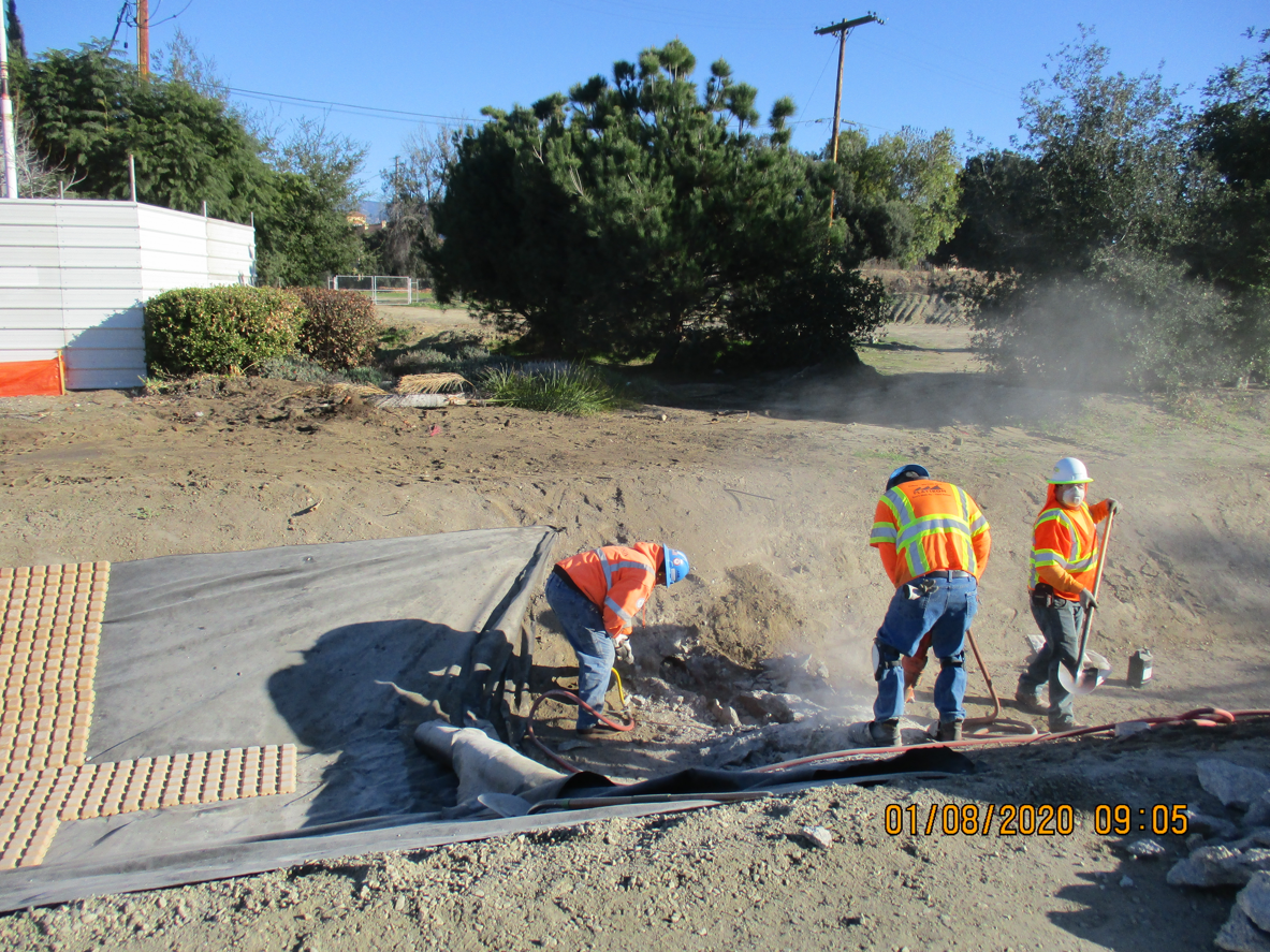 Three construction workers in safety gear dig and work with tools near a roadside curb on a dusty construction site. Trees and power lines are visible in the background.