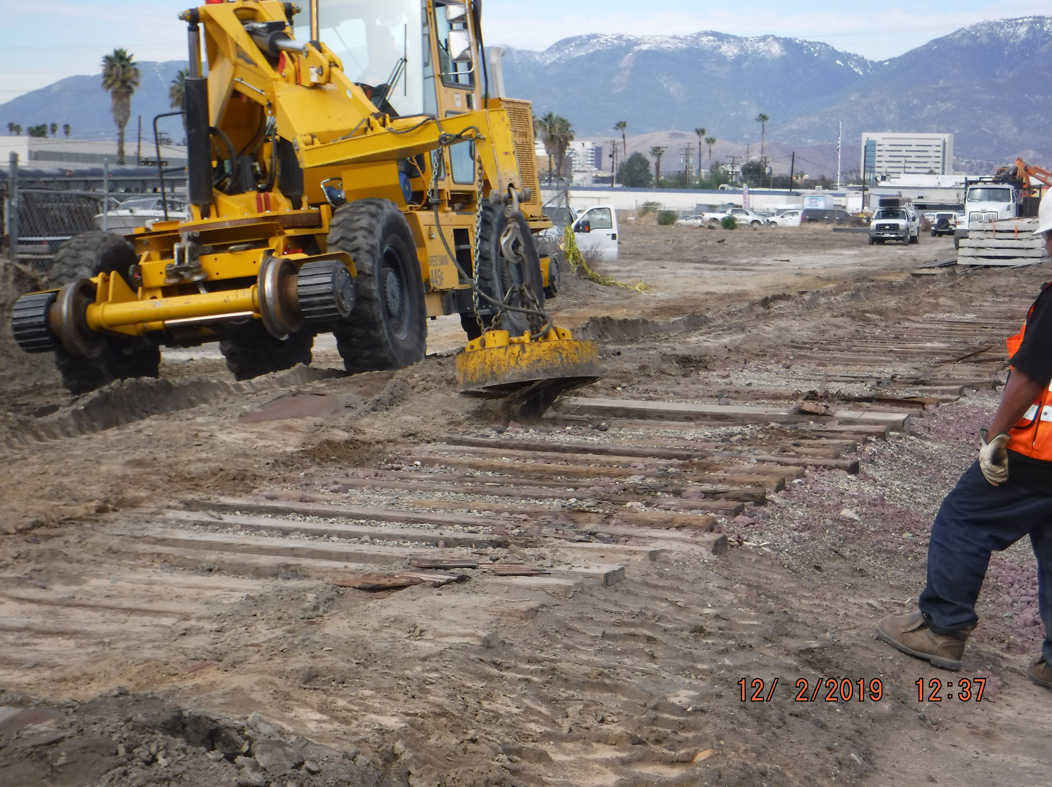 A yellow construction vehicle removes old wooden railroad ties from a dirt lot; mountains and buildings are visible in the background.