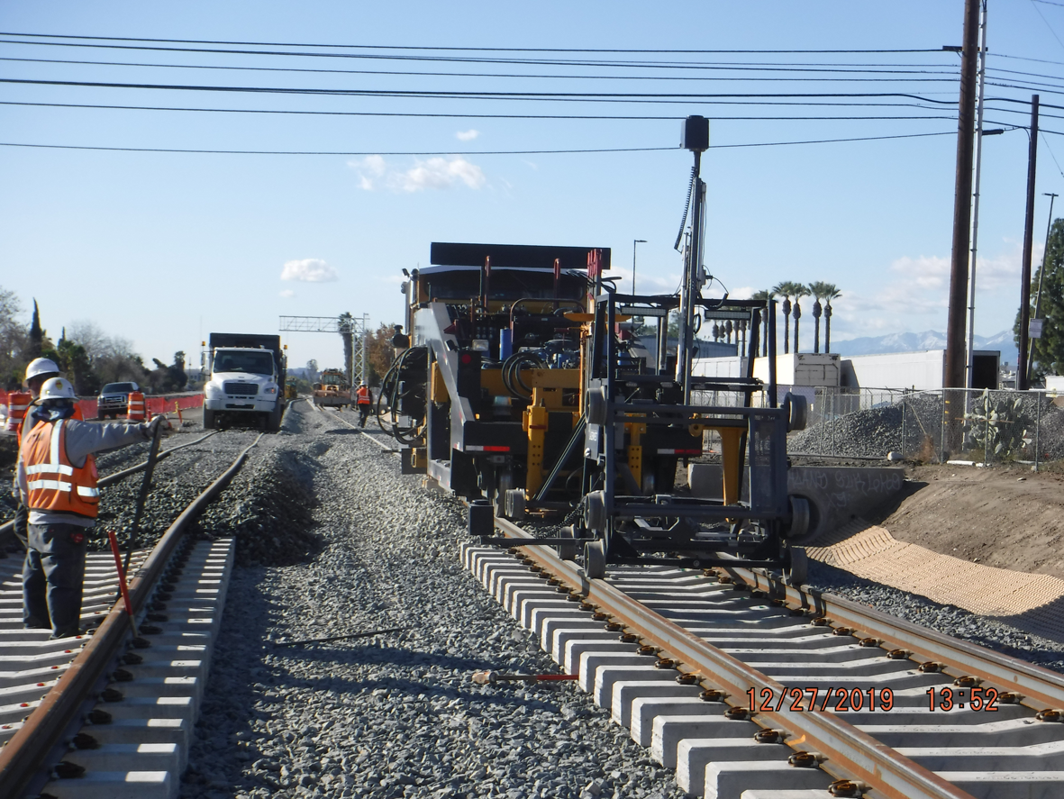Construction workers and heavy machinery work on installing new railroad tracks along a gravel bed, with vehicles and equipment visible in the background.