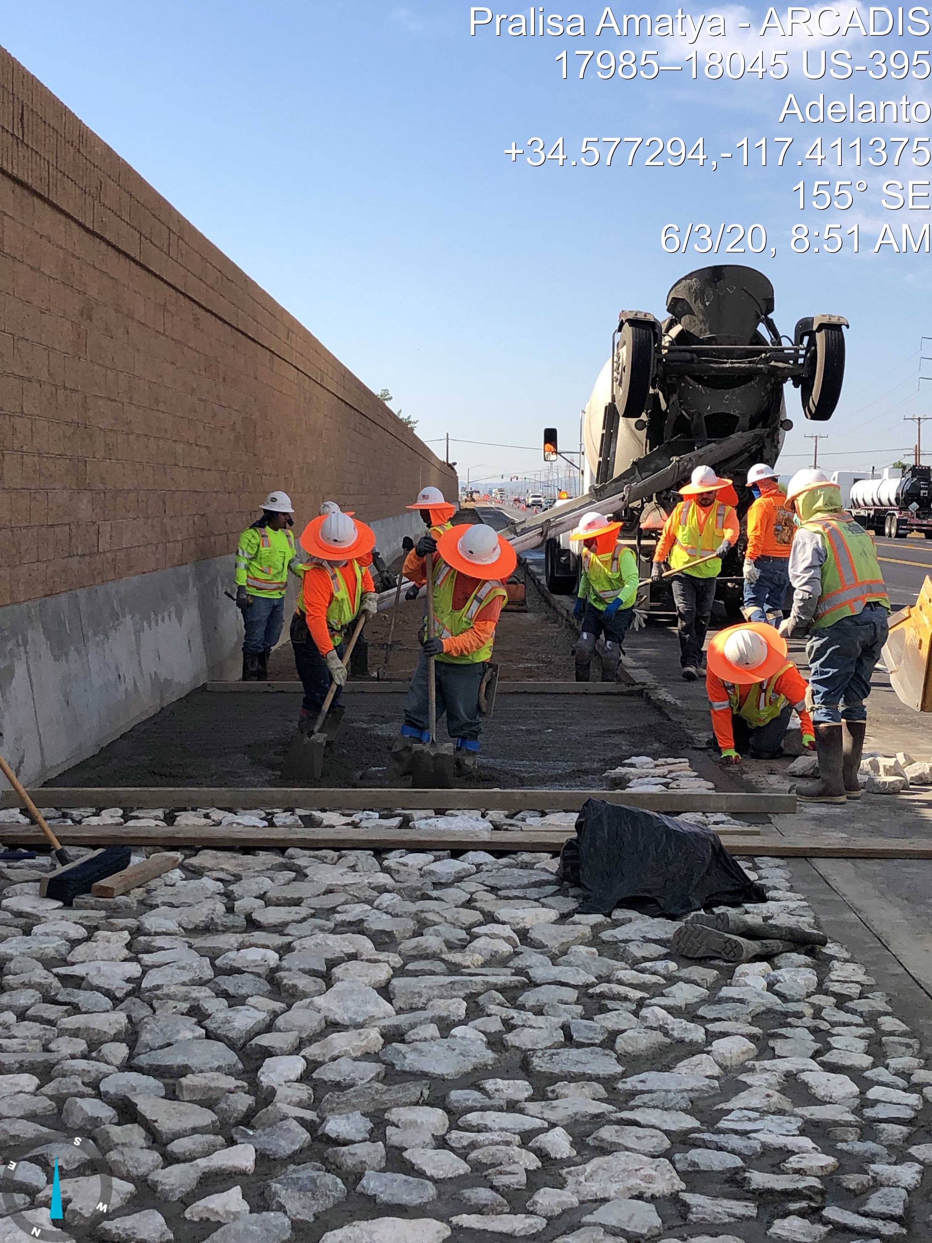 Construction workers in safety vests and helmets spread and smooth concrete on a roadway beside a cement truck, near a stone-lined drainage area and a concrete wall.
