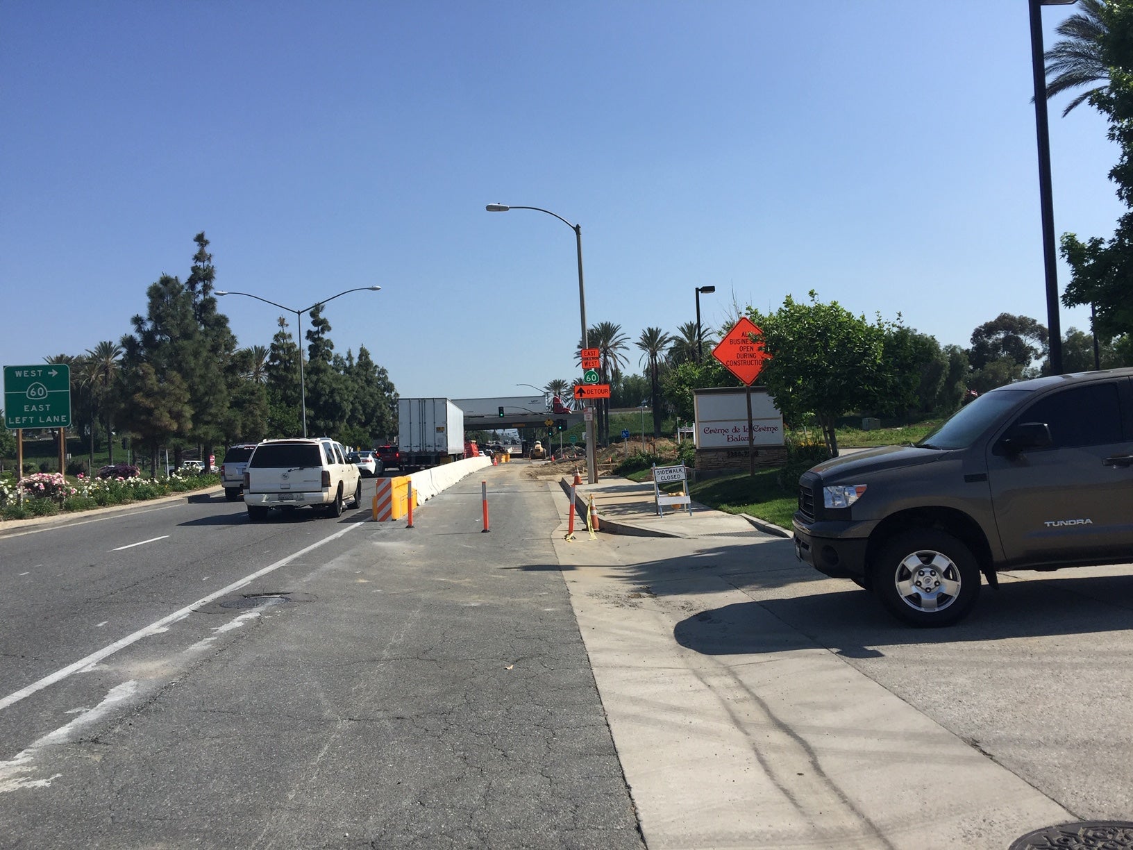 Street scene with road construction, traffic cones, work signs, and vehicles parked along the side; trees and streetlights line the road under a clear sky.