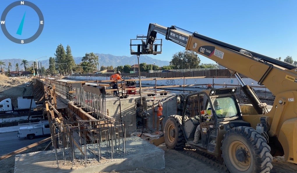 Construction workers use heavy machinery to work on a bridge structure. Steel reinforcements and concrete are visible. A clear sky and mountains are in the background.