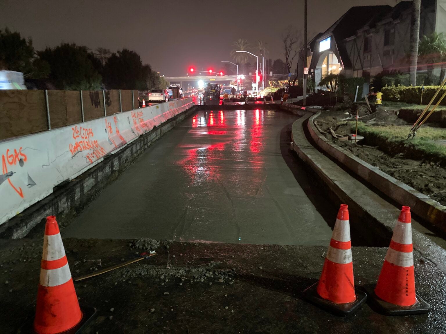 Nighttime street construction scene with fresh concrete, traffic cones in the foreground, and red lights reflecting on the wet surface.