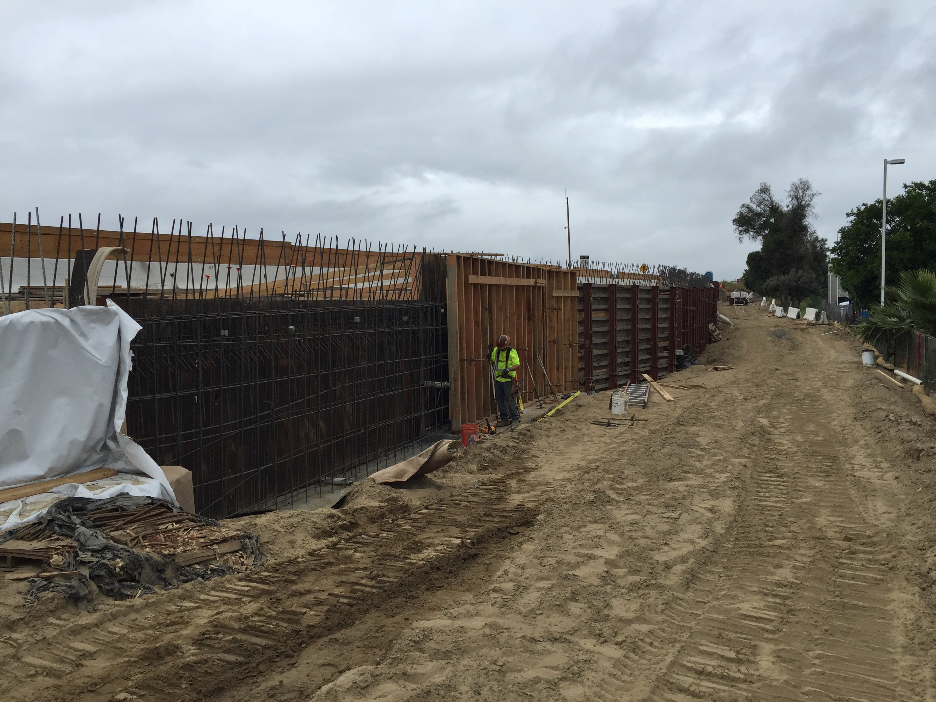 A construction worker stands by a partially built concrete wall reinforced with rebar along a dirt path, with cloudy skies overhead.