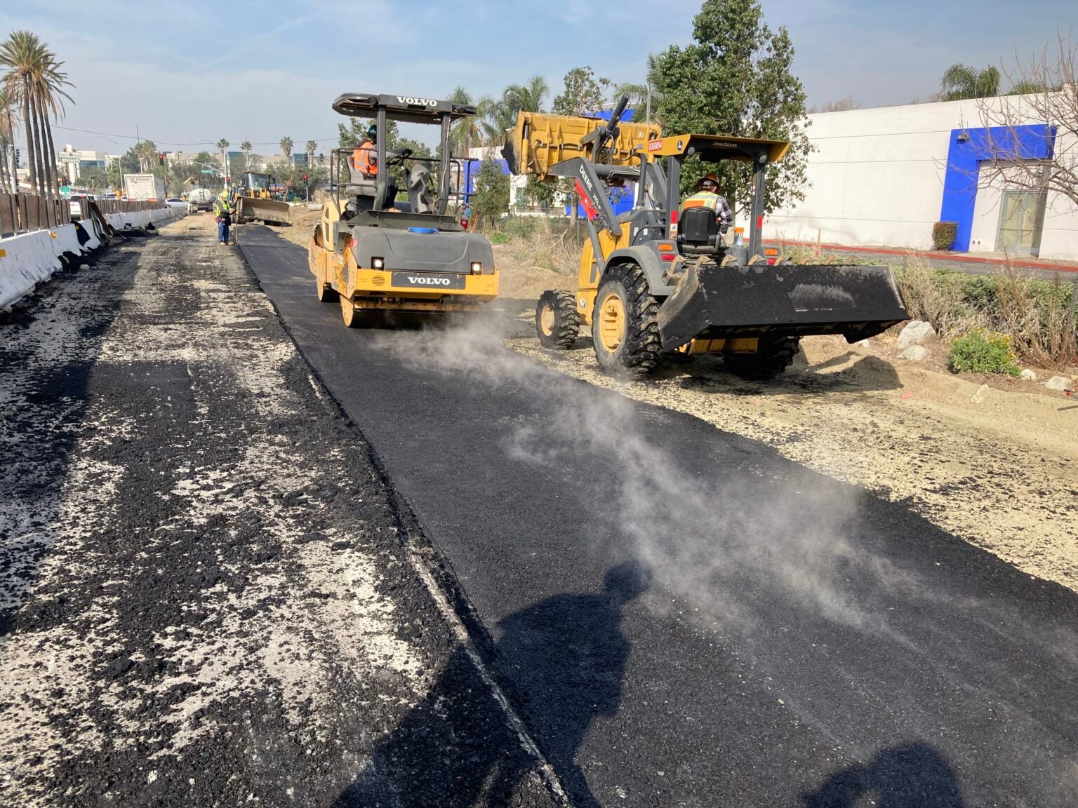 Construction vehicles are paving a road with fresh asphalt, steam rising from the surface, while workers operate the machinery under a clear sky.