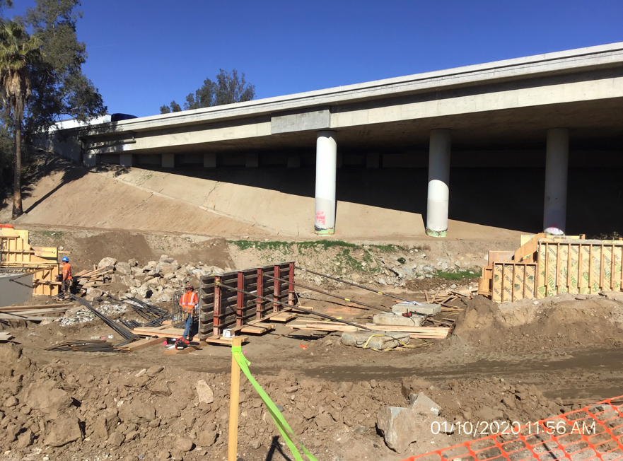 Construction workers at a work site beneath an overpass, surrounded by dirt, rocks, wooden forms, and construction materials.