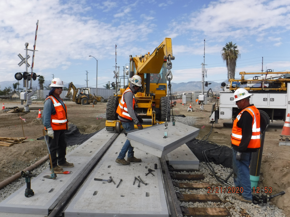 Three construction workers in safety vests install a concrete panel on a railroad crossing using a yellow crane at an outdoor worksite.