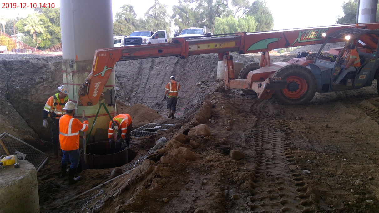 Construction workers in safety vests and helmets work around a column base near a telescopic handler at a dirt excavation site under an overpass.
