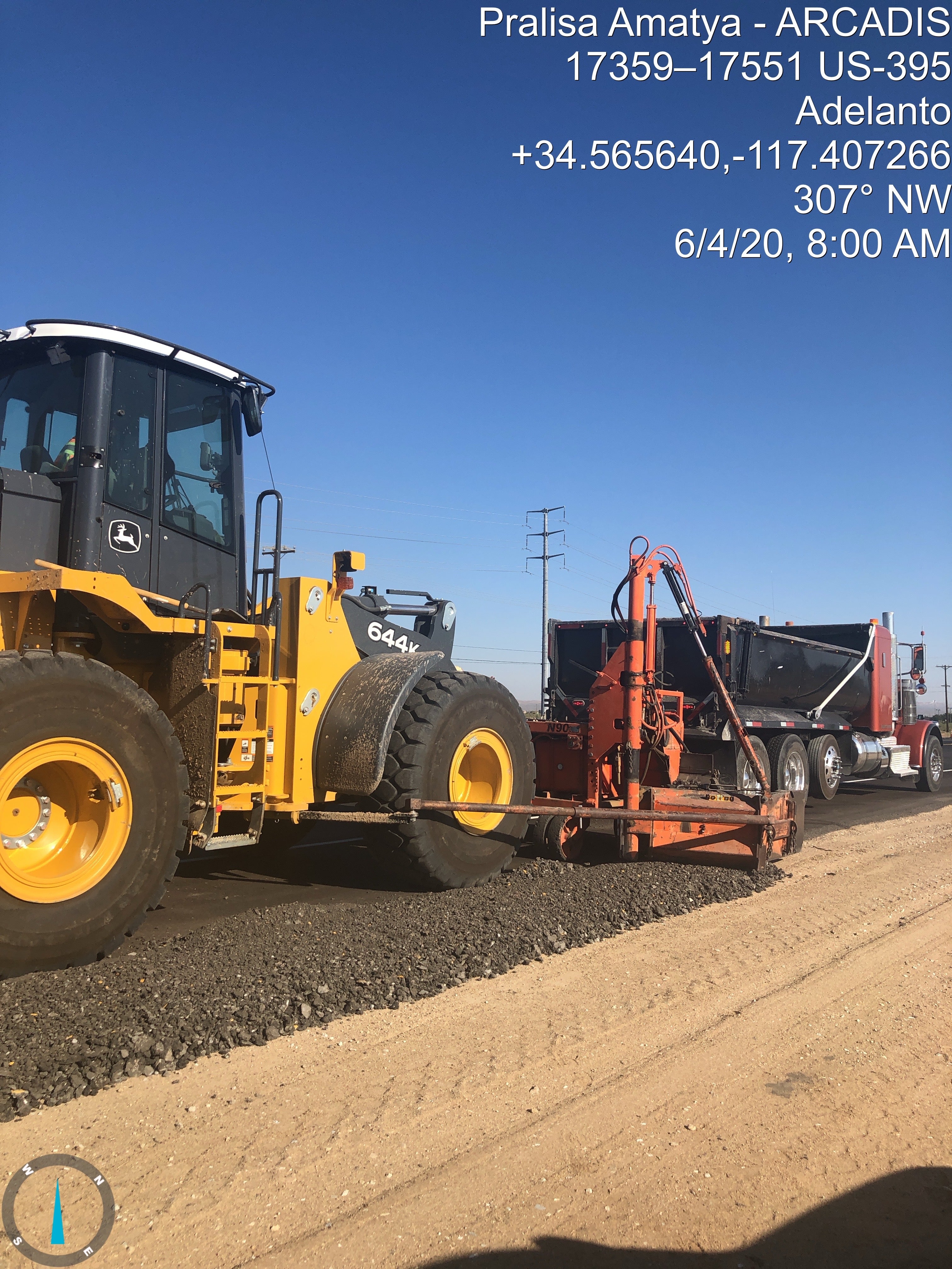 A yellow construction vehicle and a red truck working on a gravel road under a clear blue sky, with location and date details overlaid at the top right.