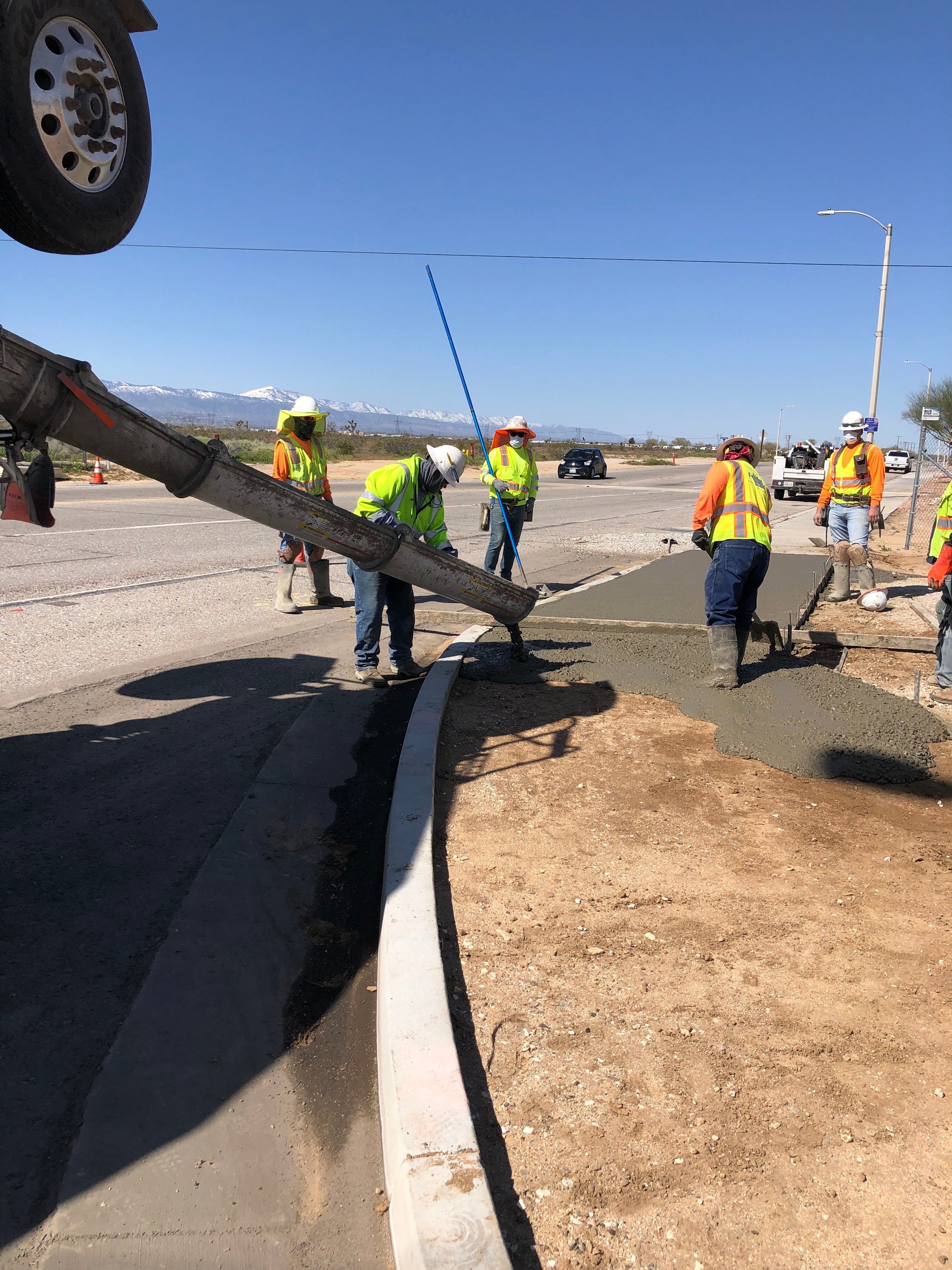 Construction workers pour and spread concrete to form a sidewalk along a roadside on a sunny day.