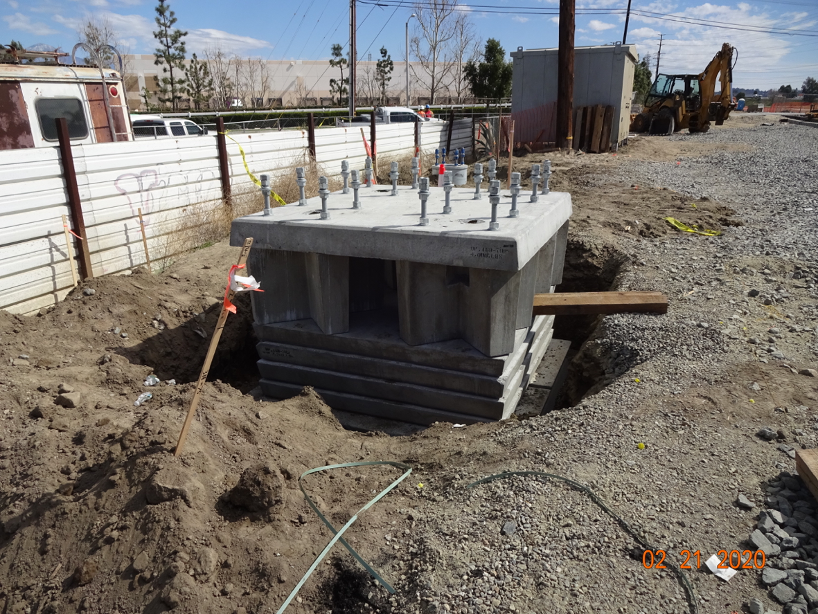 A large concrete foundation with anchor bolts is partially buried in a dirt construction site, surrounded by equipment and materials.