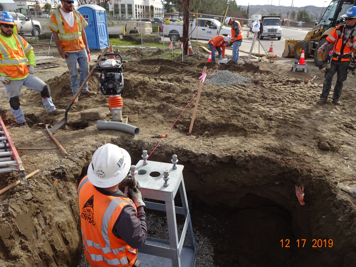 Construction workers in safety vests and helmets install utilities and operate machinery at an outdoor worksite with dirt, equipment, and traffic cones visible.