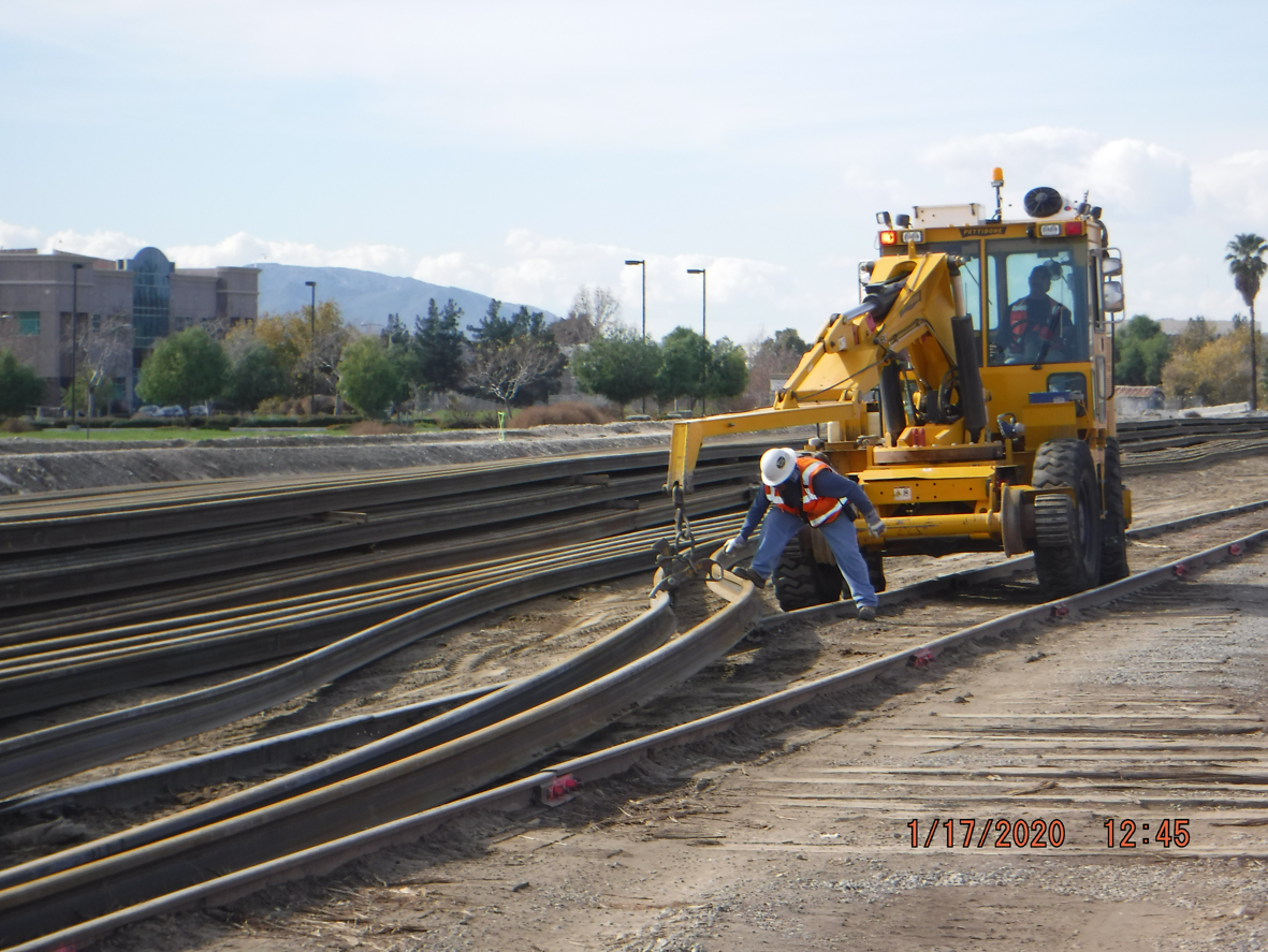 A worker adjusts a section of railroad track while another operates a yellow maintenance vehicle on a rail yard during daytime.