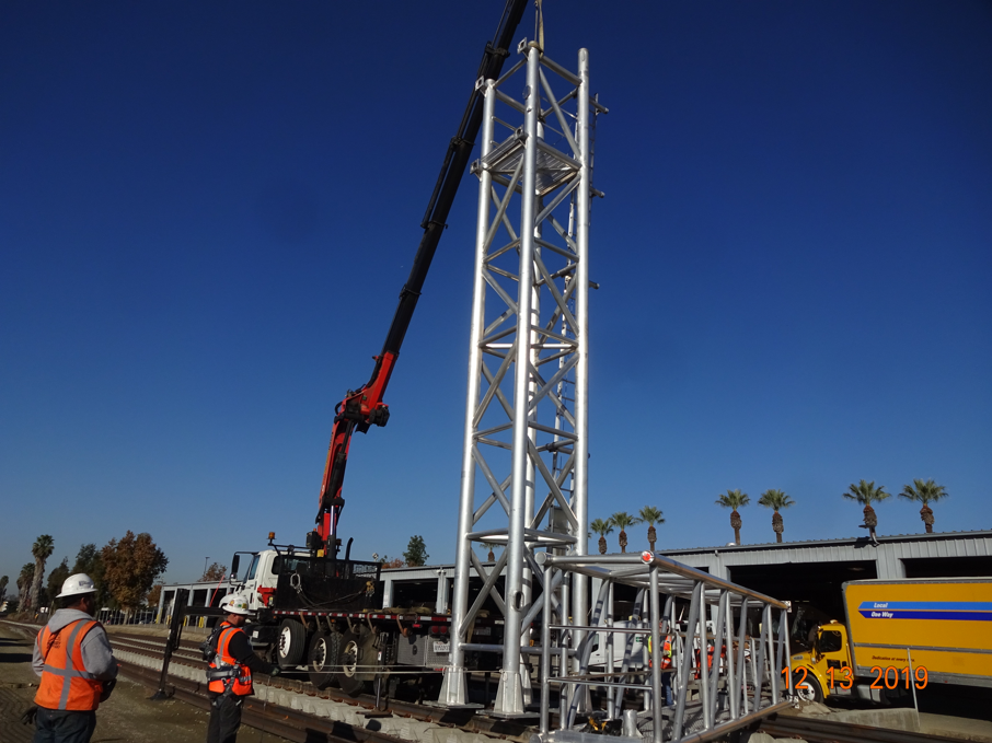 A crane lifts a large metal tower structure near train tracks as workers in safety gear supervise the operation on a sunny day.
