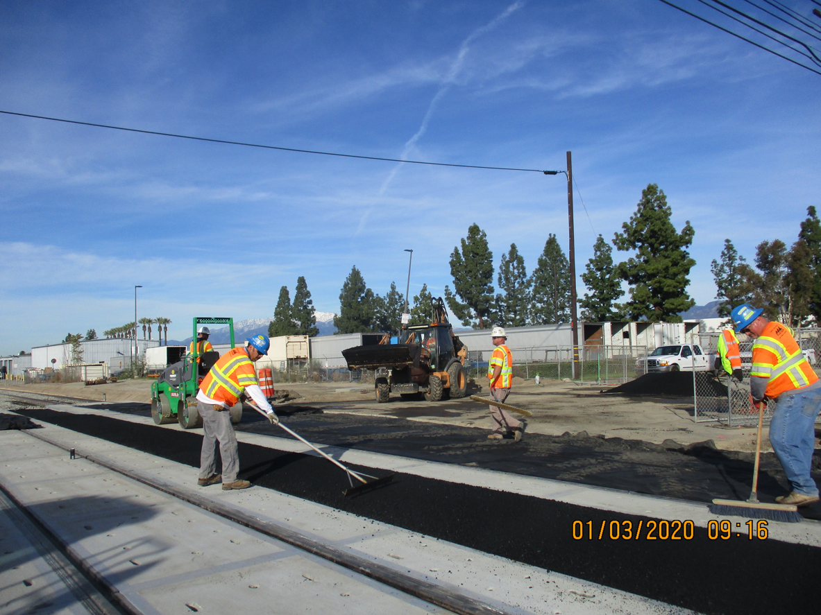 Construction workers in safety gear spread asphalt on a road near rail tracks using tools and machinery on a clear day.