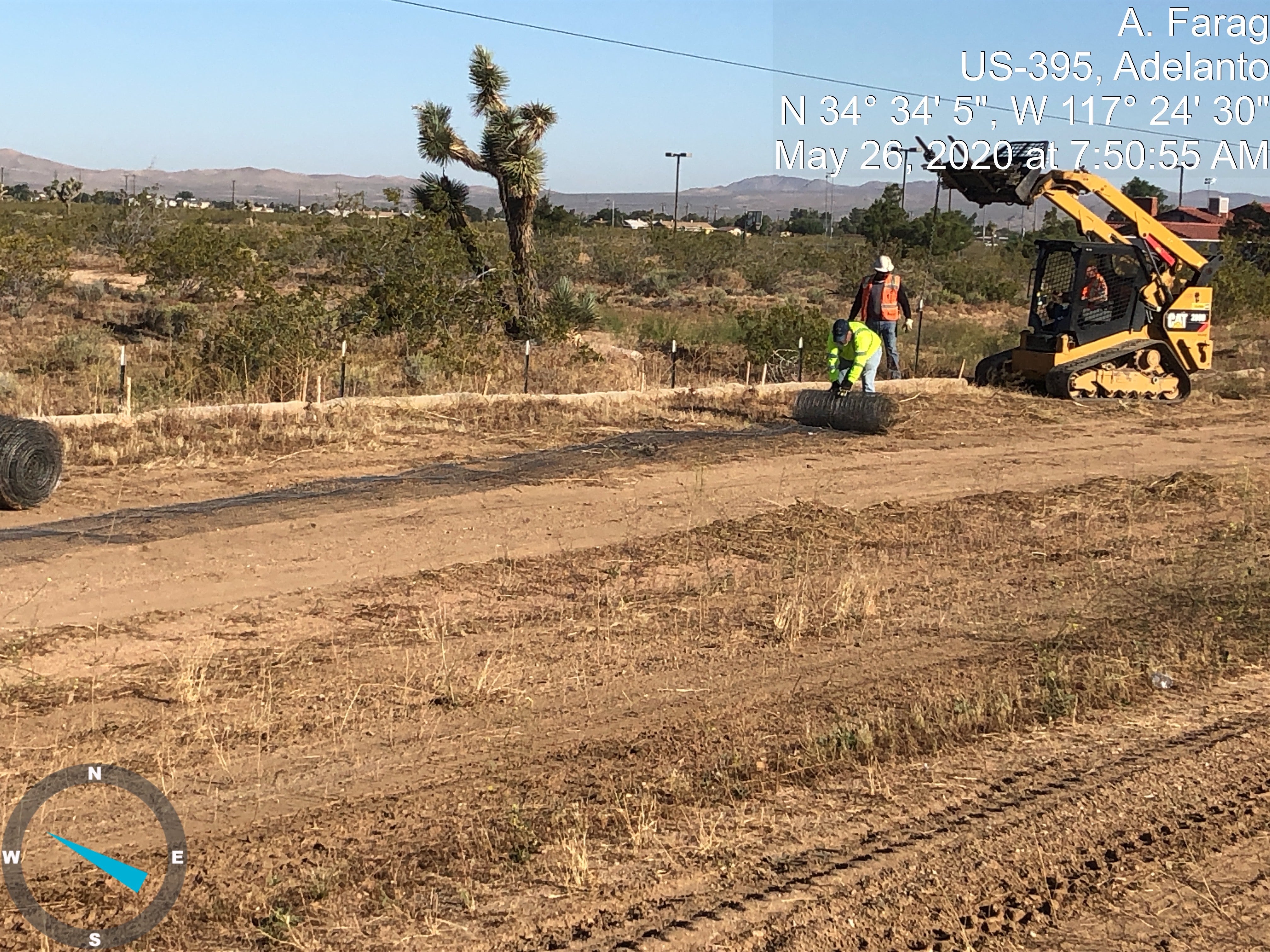 Two construction workers move a roll of wire fence on a dirt road in a desert area, with a small loader and sparse vegetation in the background.
