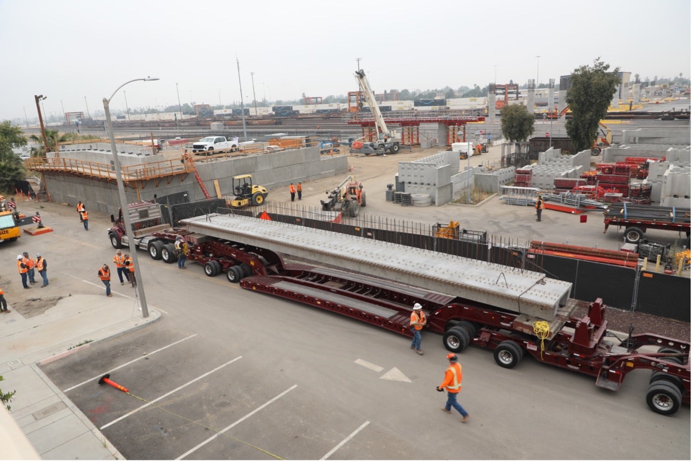 A large steel beam is transported on a multi-axle trailer through a construction site, with workers in orange safety vests guiding the process.