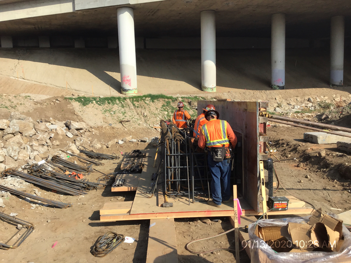 Two construction workers in safety gear assemble rebar under a highway overpass at a bridge construction site. Loose rebar and tools are scattered on the ground nearby.