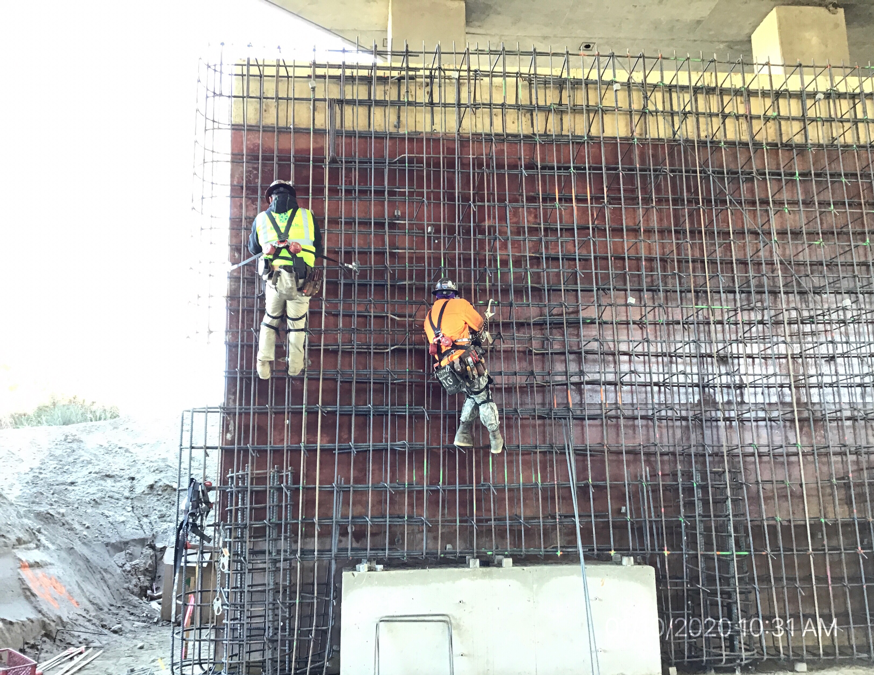 Two construction workers in safety gear secure rebar to a vertical structure at a building site, suspended by harnesses under a concrete overpass.