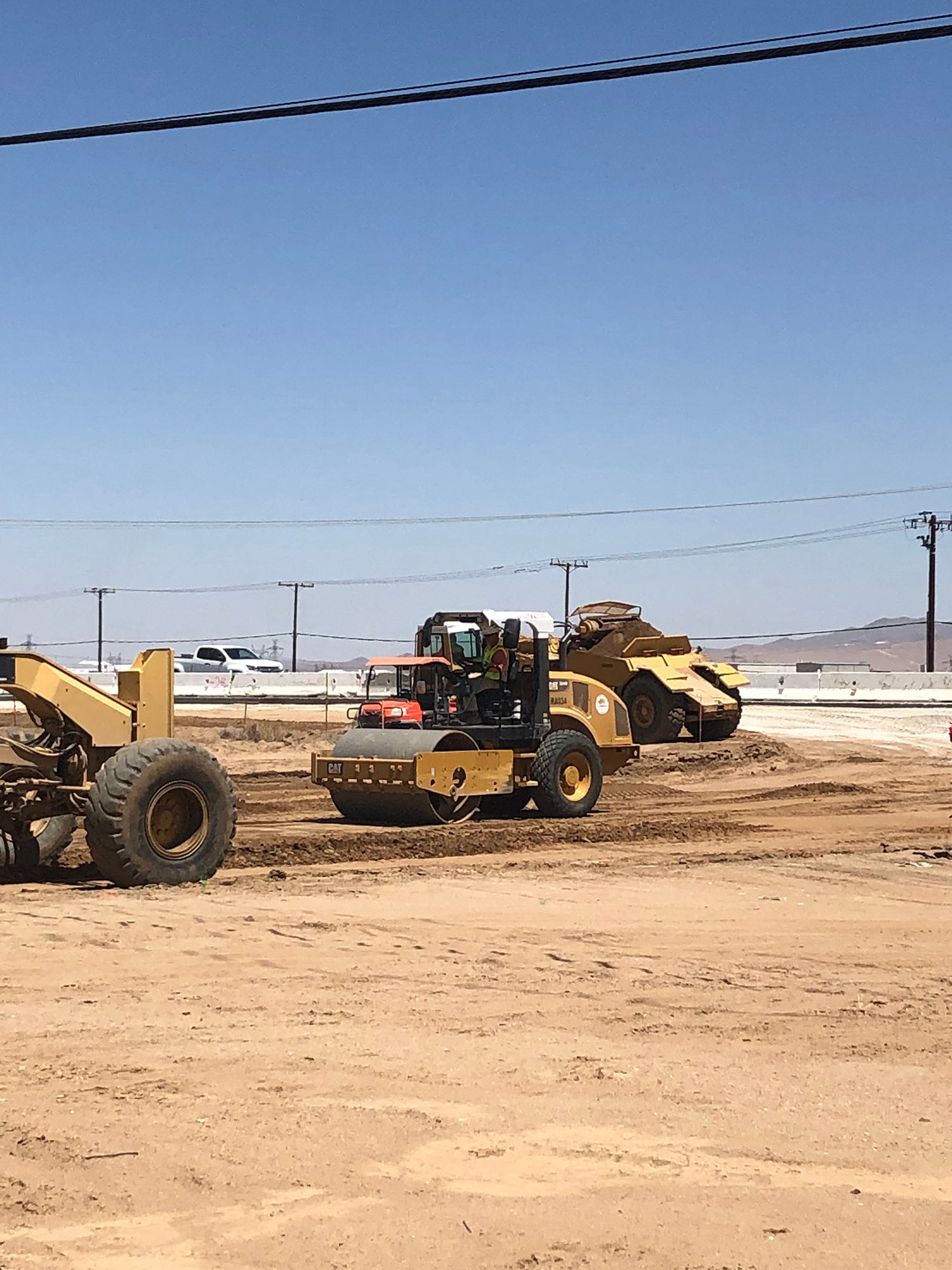 Construction vehicles working on a dirt lot under clear blue sky, with utility poles and a road visible in the background.