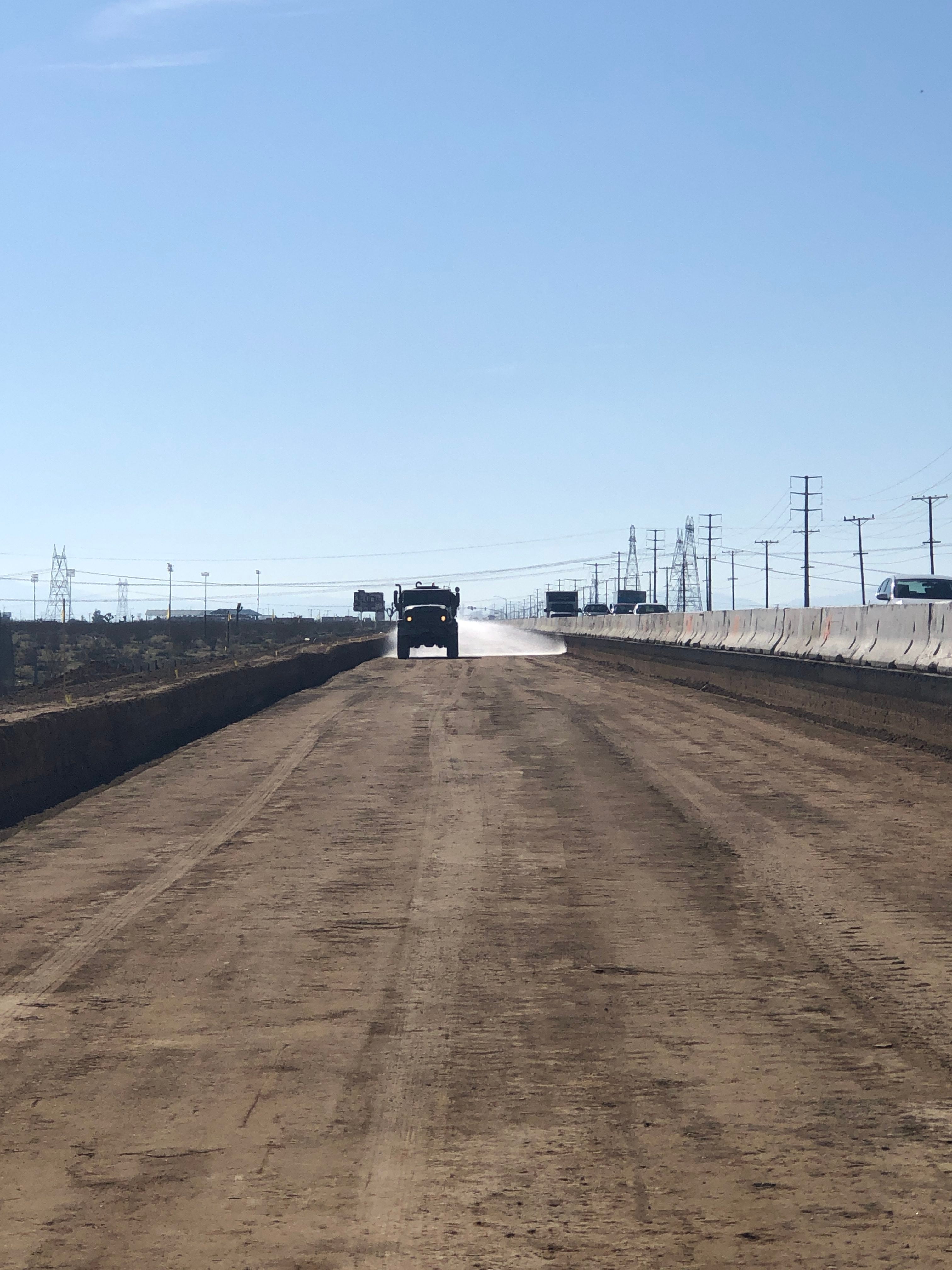 A construction vehicle drives on a wide dirt road with concrete barriers on one side and utility poles in the background under a clear sky.