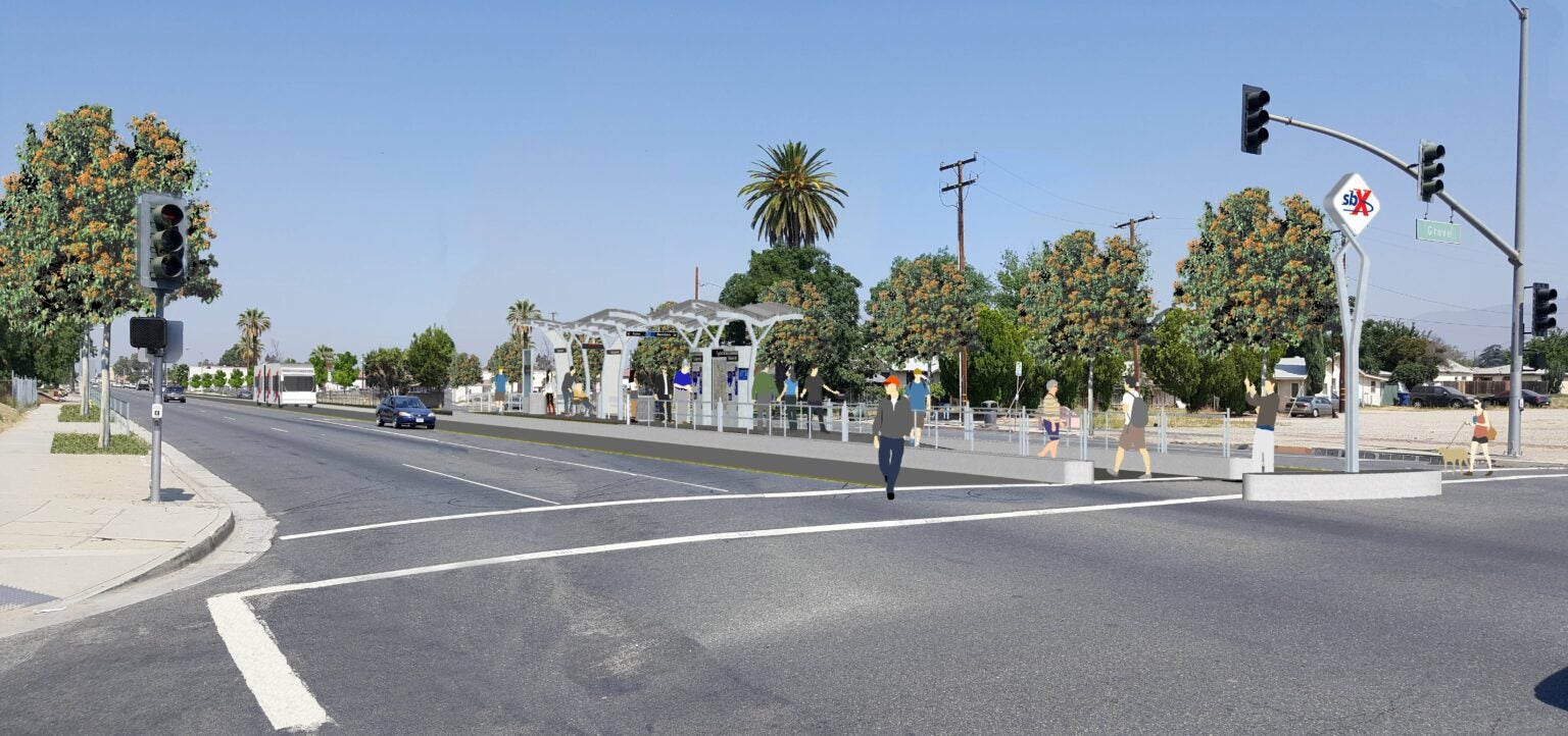 A street view with a crosswalk, traffic lights, a car driving, and people walking near a shaded bus stop or transit station with trees and signage in the background.