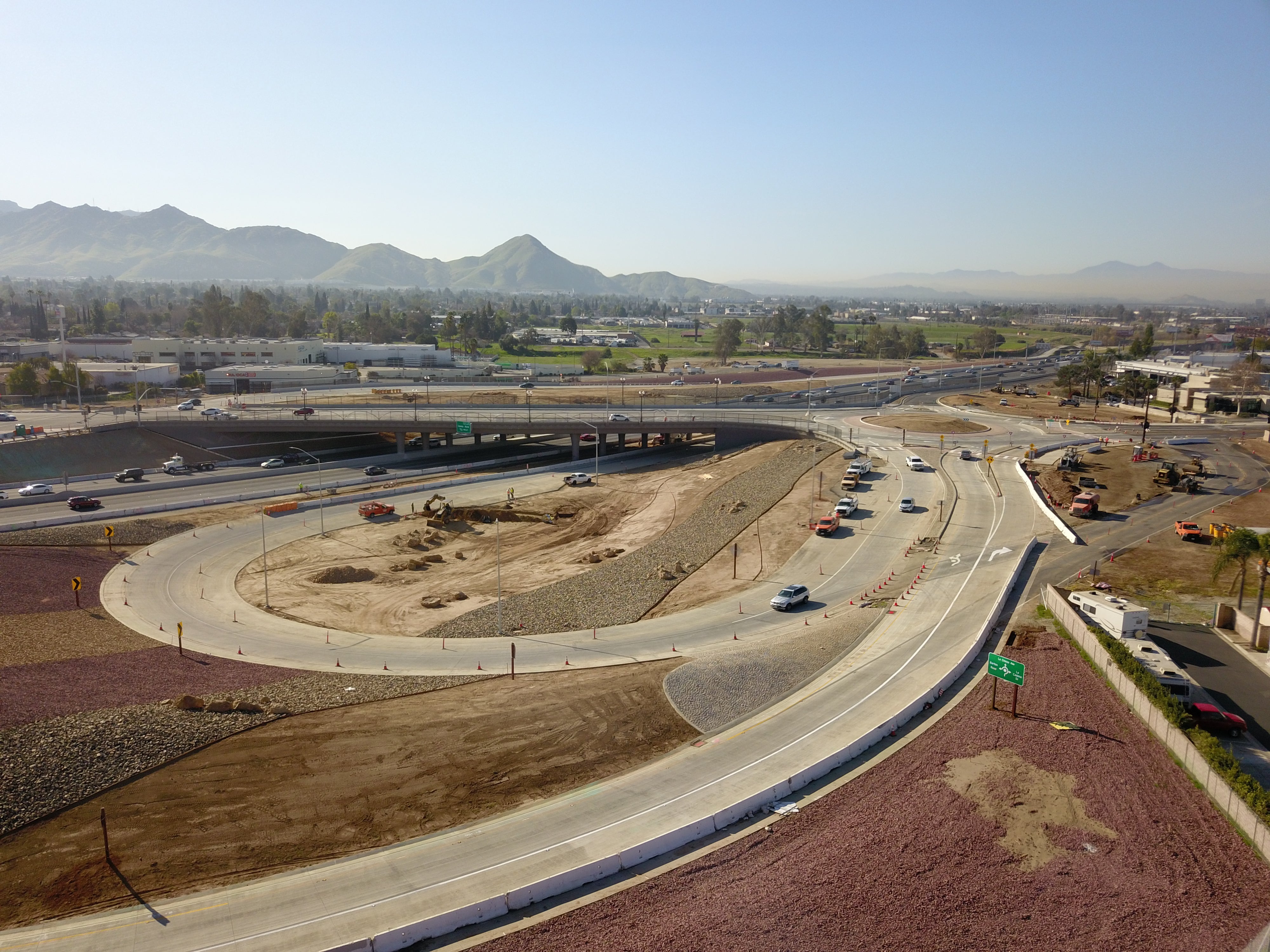 Aerial view of a highway interchange under construction with multiple lanes, vehicles, construction equipment, and surrounding landscape with mountains in the distance.