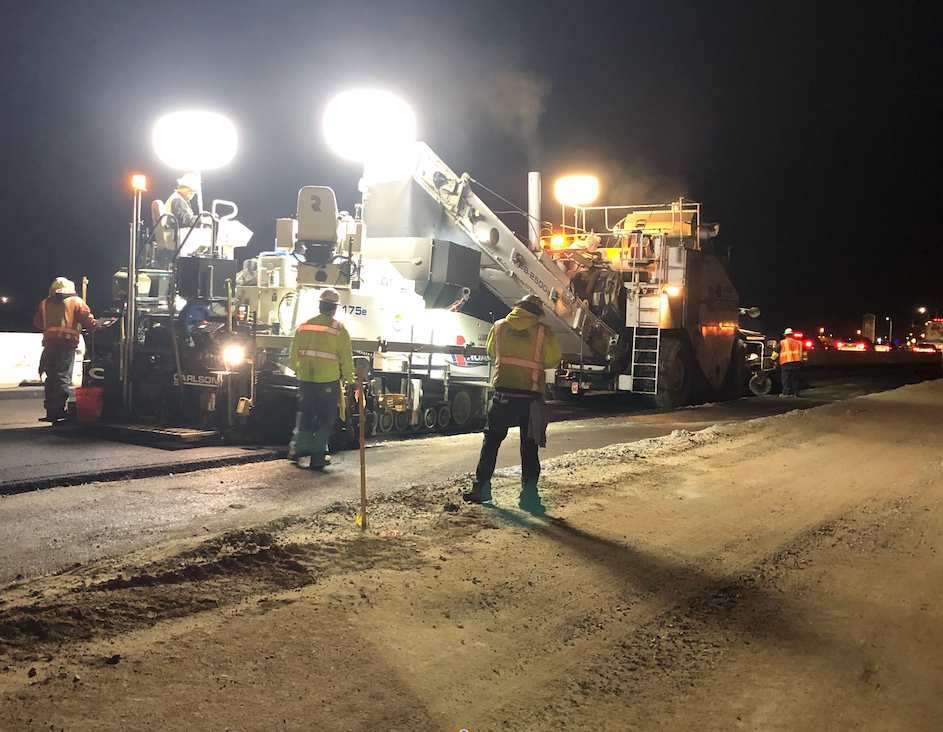 Road construction workers operate heavy machinery under bright lights at night, resurfacing a section of highway with vehicles visible in the background.