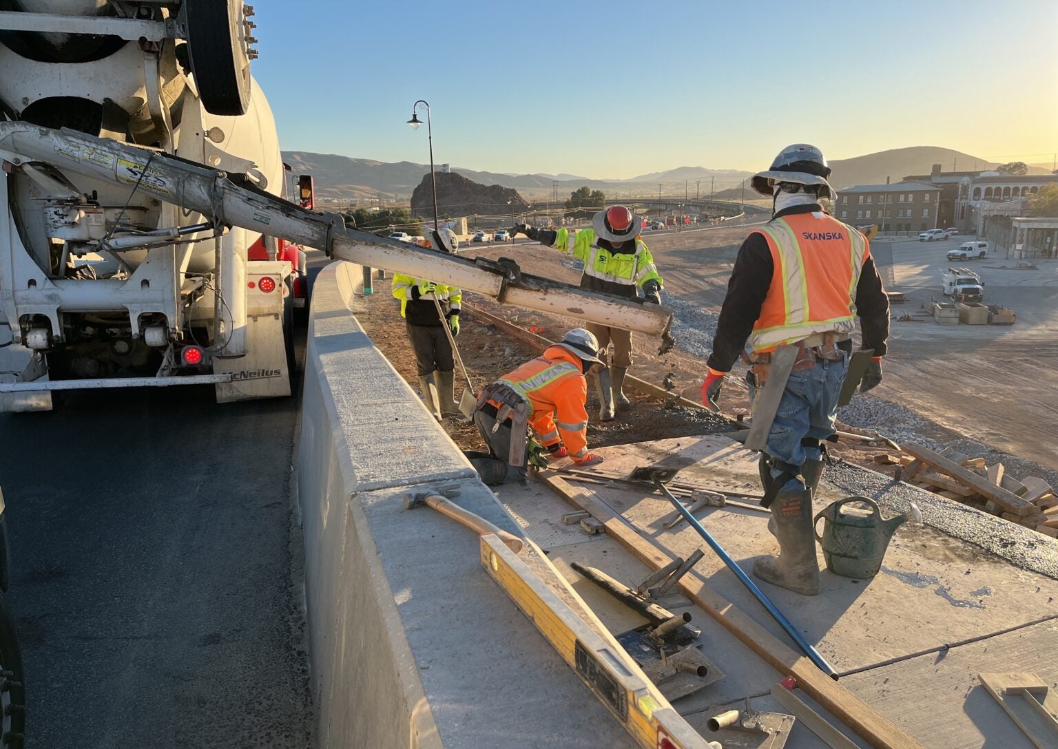 Construction workers pour concrete from a truck onto a bridge under construction at sunrise, wearing safety gear and high-visibility clothing.