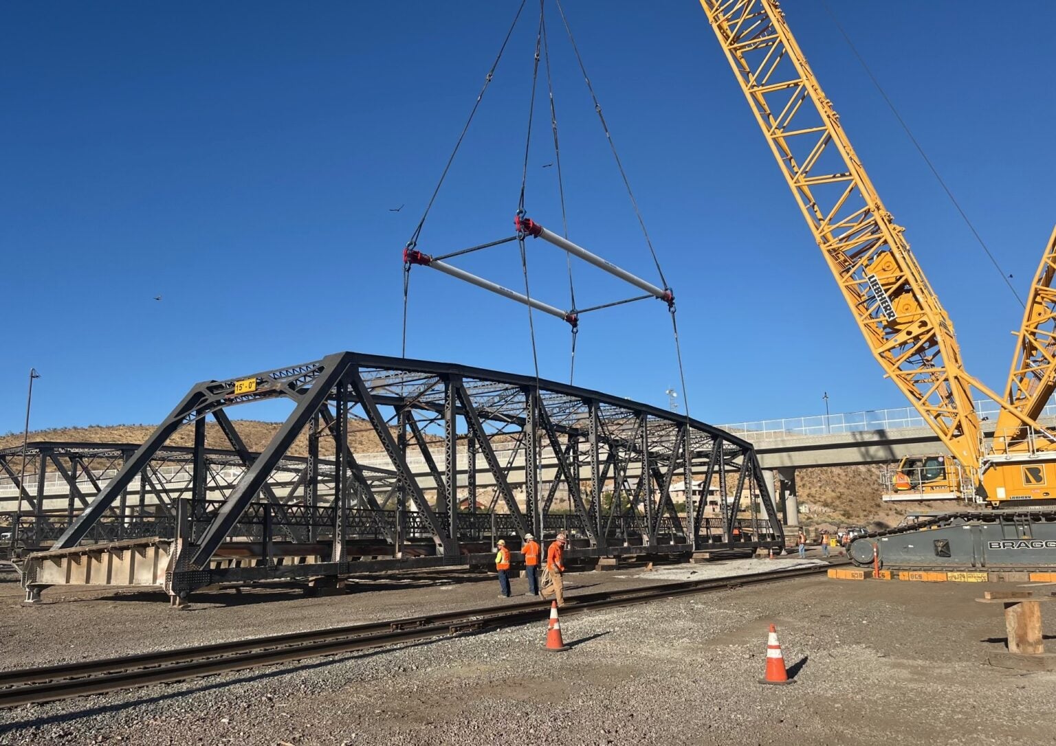 A crane lifts a large steel bridge section over railway tracks while construction workers in safety gear supervise the operation.