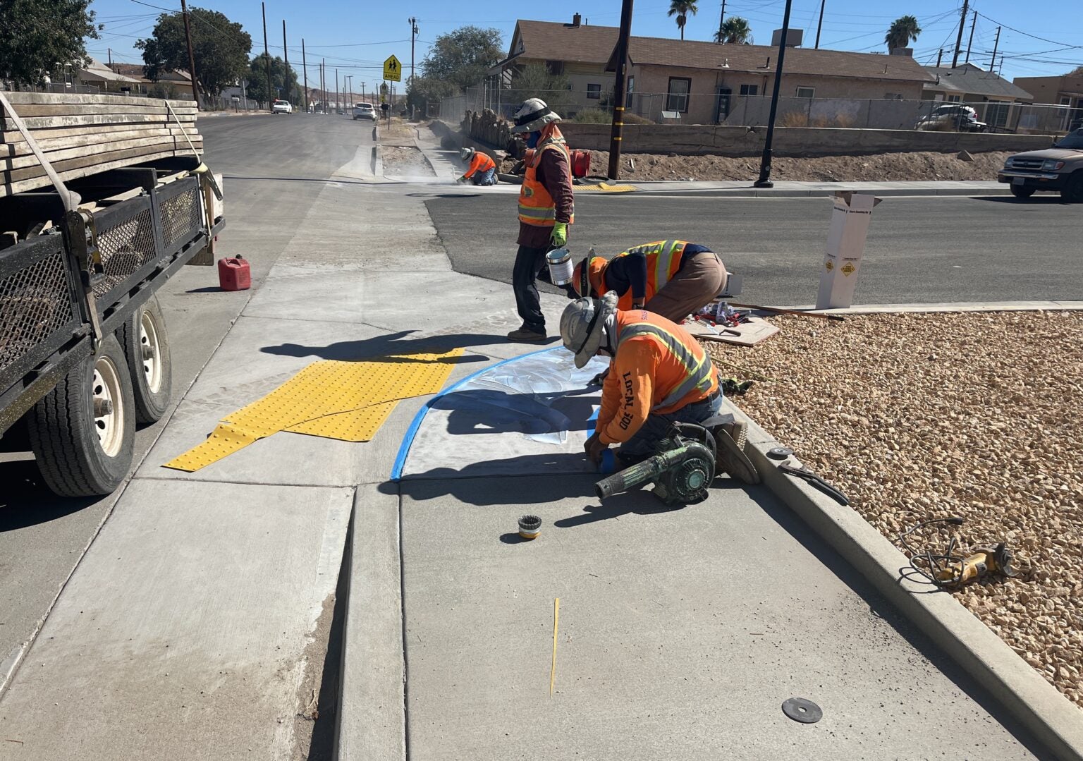 Three construction workers install and mark pavement near a curb while wearing safety gear. A trailer and construction equipment are visible nearby.