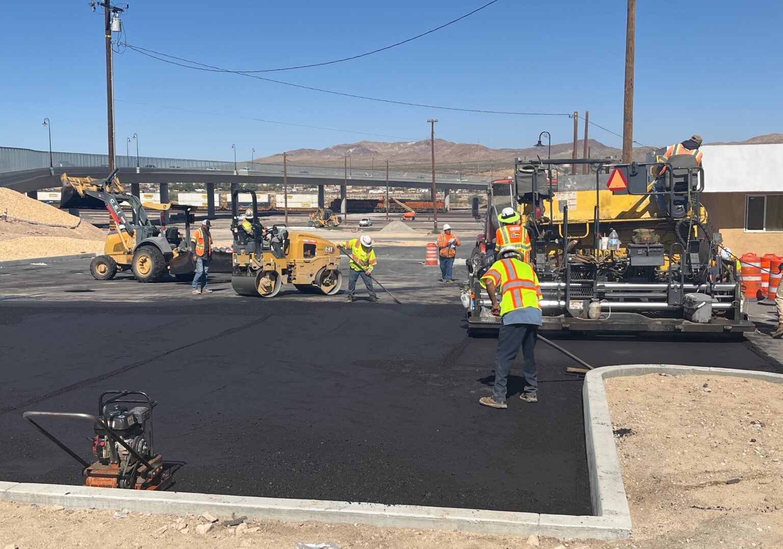 Construction workers operate heavy machinery and spread asphalt while paving a parking lot under clear skies in an industrial area.