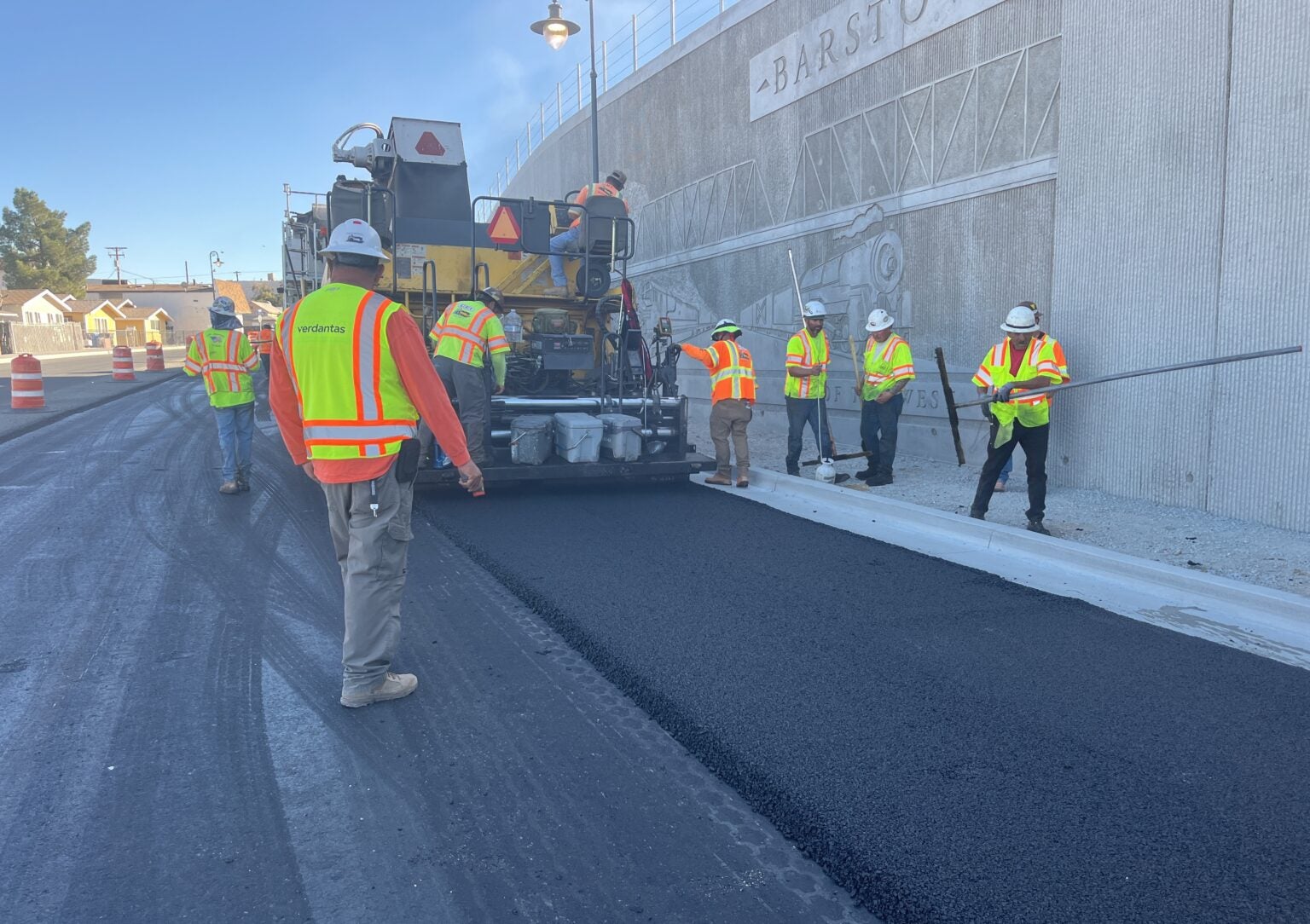 Construction workers in safety vests operate machinery and lay fresh asphalt on a road next to a concrete wall with a mural.