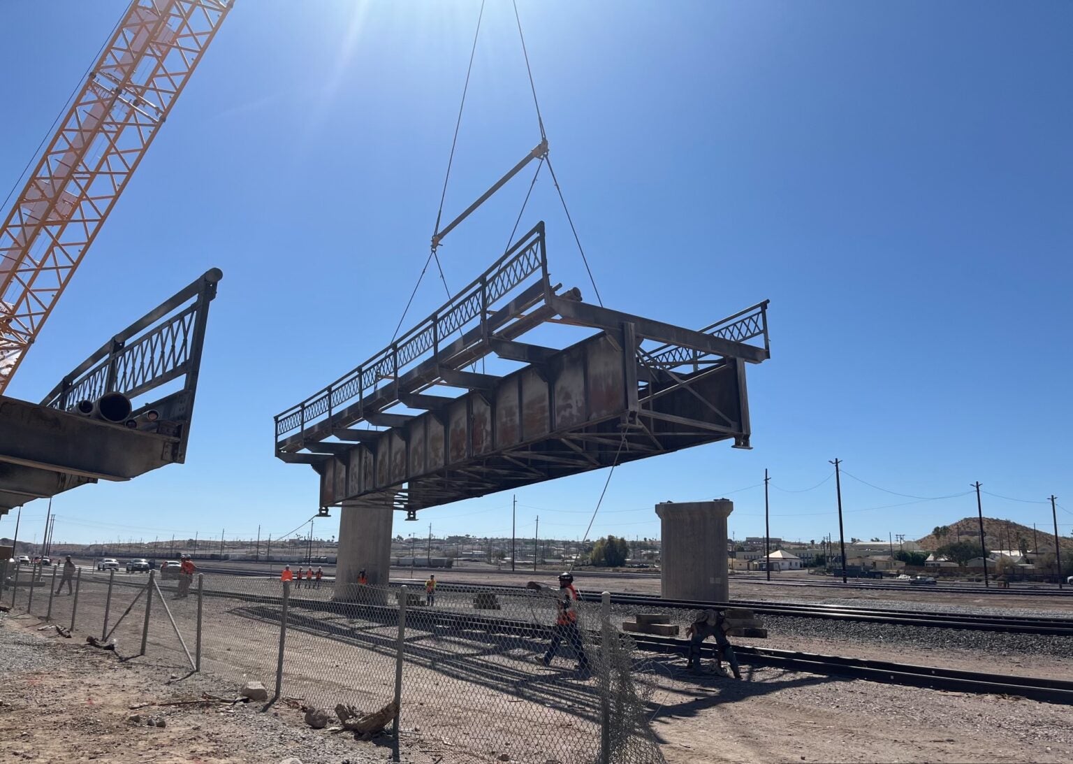 A large crane lifts a metal bridge section onto concrete supports over railway tracks in a construction site under a clear blue sky.