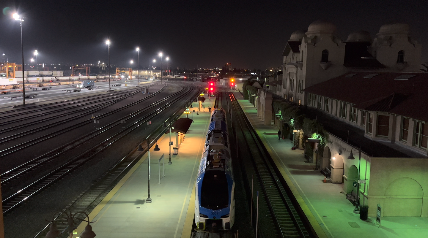 A passenger train is parked at a well-lit station platform at night, with multiple tracks and a historic building nearby.