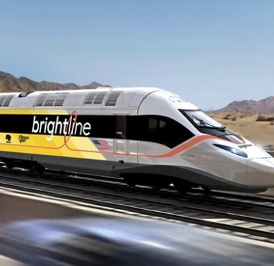 A Brightline high-speed train travels on tracks through a desert landscape with mountains in the background and blurred cars nearby.